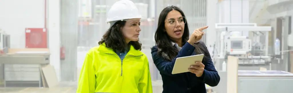 Two women in a factory, one in a high-visibility jacket and hard hat, the other—a facility manager—in business attire holding a tablet and pointing. Machinery and industrial equipment are visible in the background, likely being monitored via facilities management software.