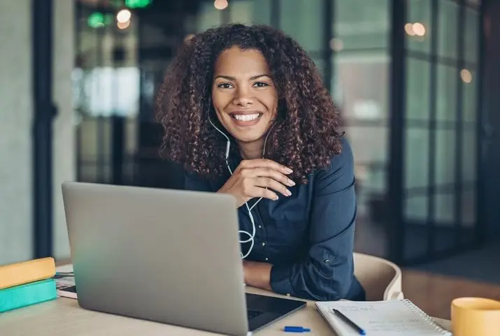 A cheerful woman with headphones is seated at a desk, engaged with her laptop and smiling.