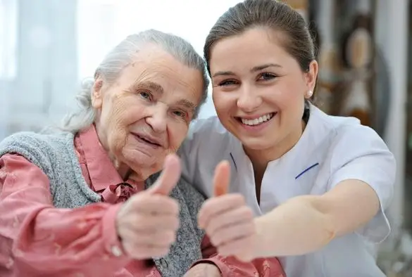 A nurse smiles and gives a thumbs up to an elderly woman, conveying encouragement and support in a healthcare setting.