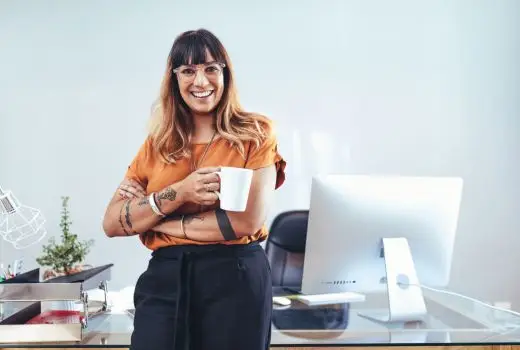 A woman stands by a desk, holding a coffee cup, with a focused expression on her face.