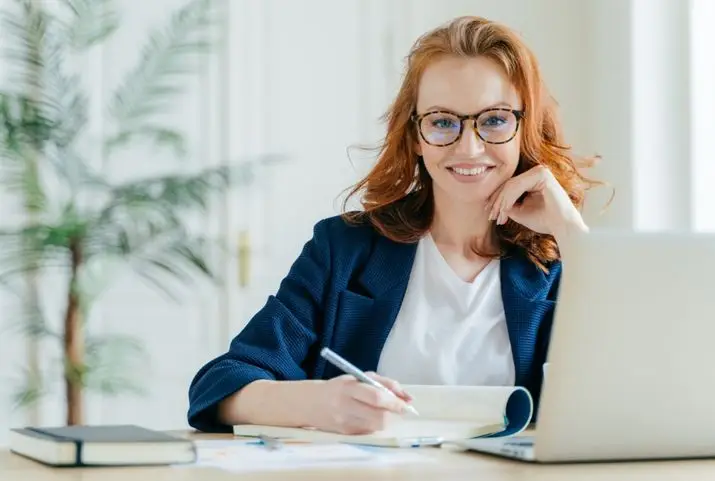 A woman with glasses is seated at her desk, engaged with her laptop in a tidy workspace.
