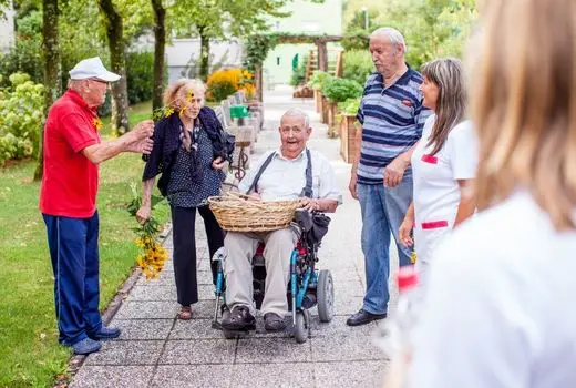 Man in wheelchair holds basket. He is surrounded by neighbours at his retirement community.