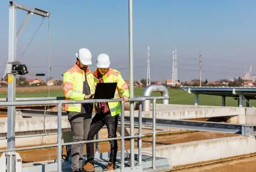 Two workers in hi vis vests and hard hats look at laptop on substation site
